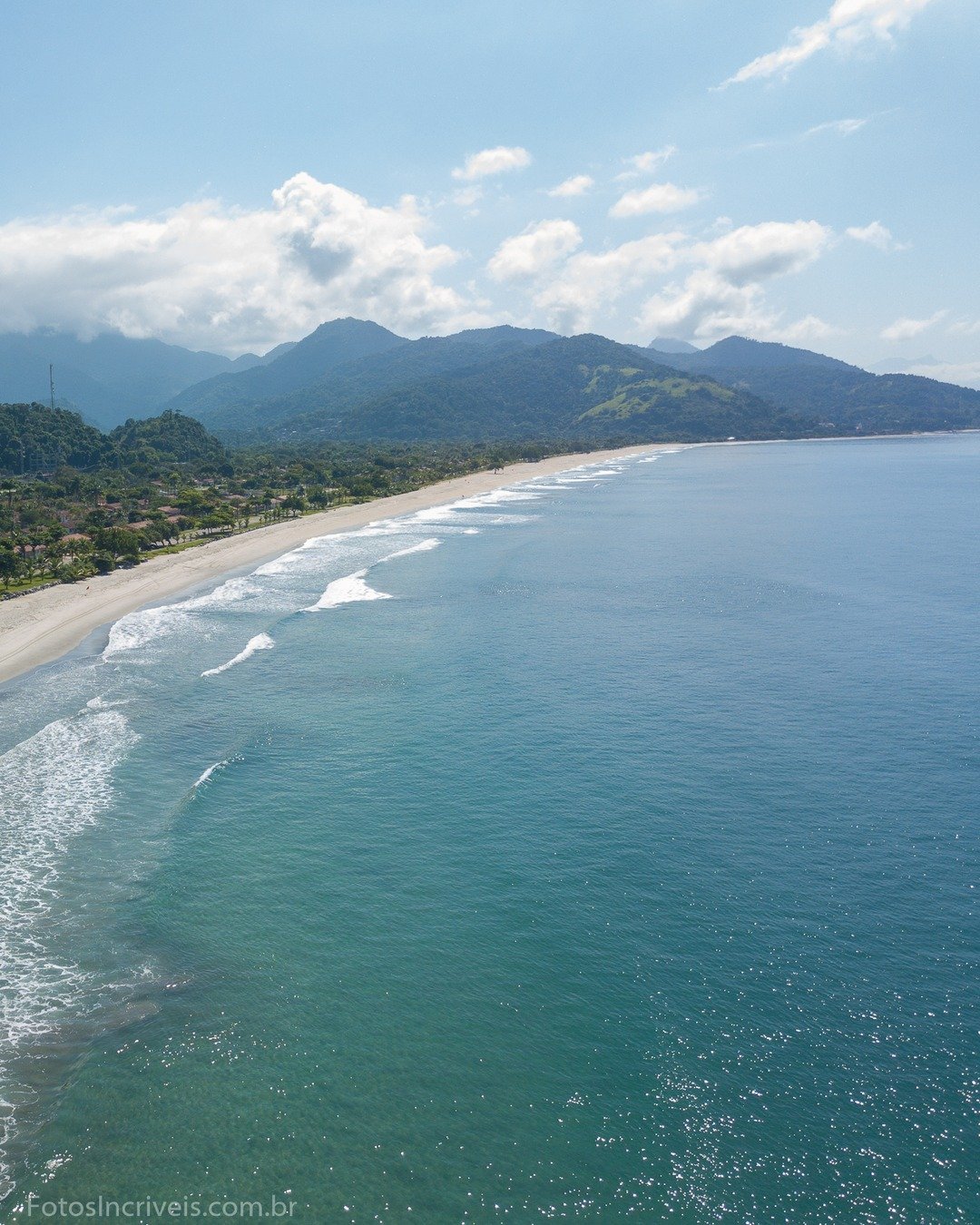 Vista aérea de uma praia com mar azul-turquesa, faixa de areia, vegetação e montanhas ao fundo.