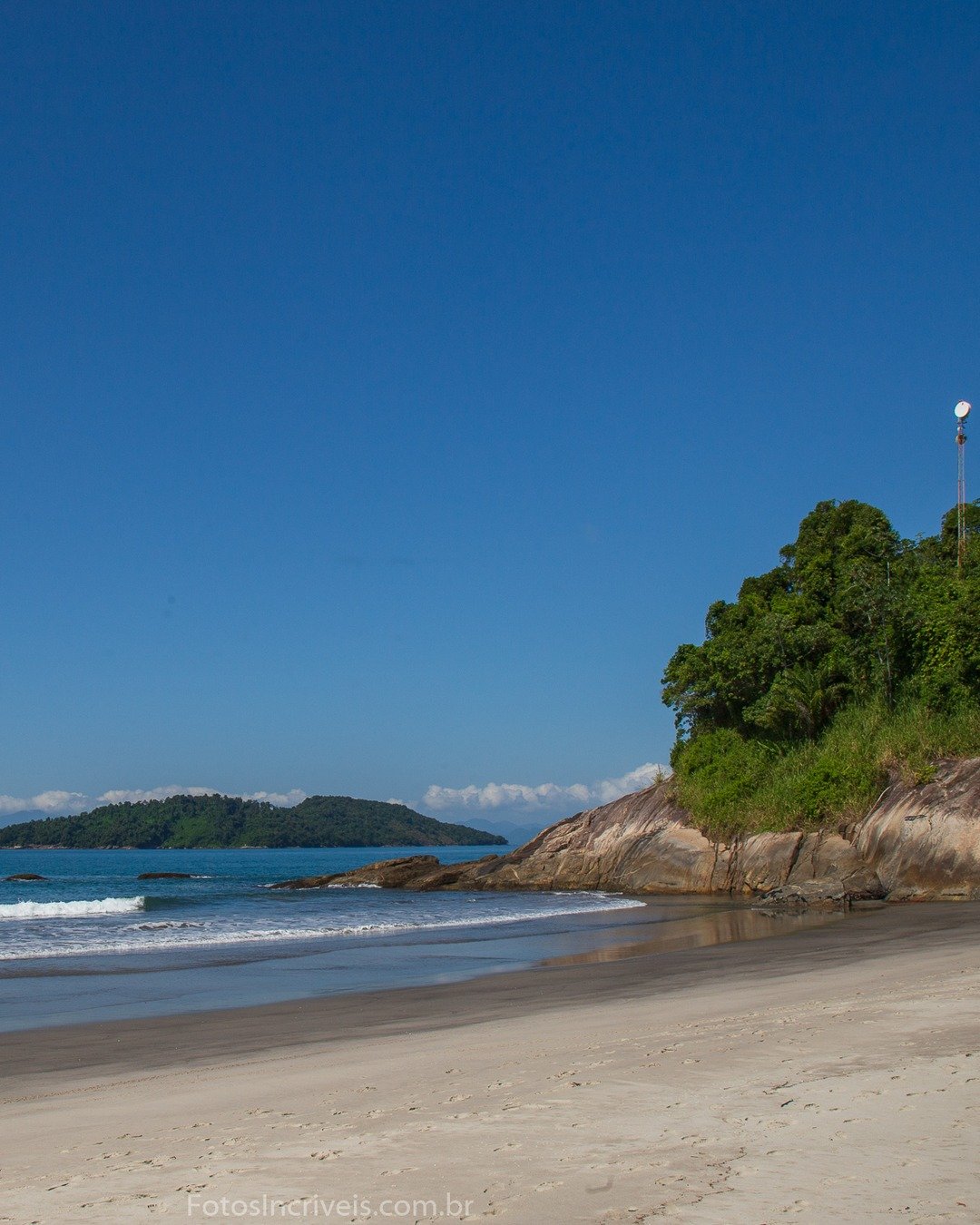 Praia com mar calmo, ondas quebrando na areia, formação rochosa com vegetação à direita e ilha ao fundo sob céu azul.