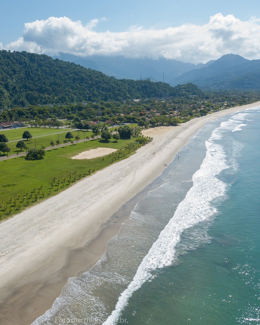 Vista aérea de uma longa faixa de praia com mar de ondas brancas à direita, areia clara, área verde gramada com árvores, estrada e montanhas cobertas de floresta ao fundo sob céu com nuvens.