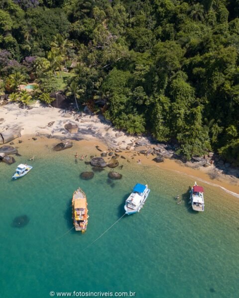 Vista aérea de uma pequena enseada com praia de areia, pedras na água, mata atlântica densa ao fundo e barcos ancorados nas águas turquesa.