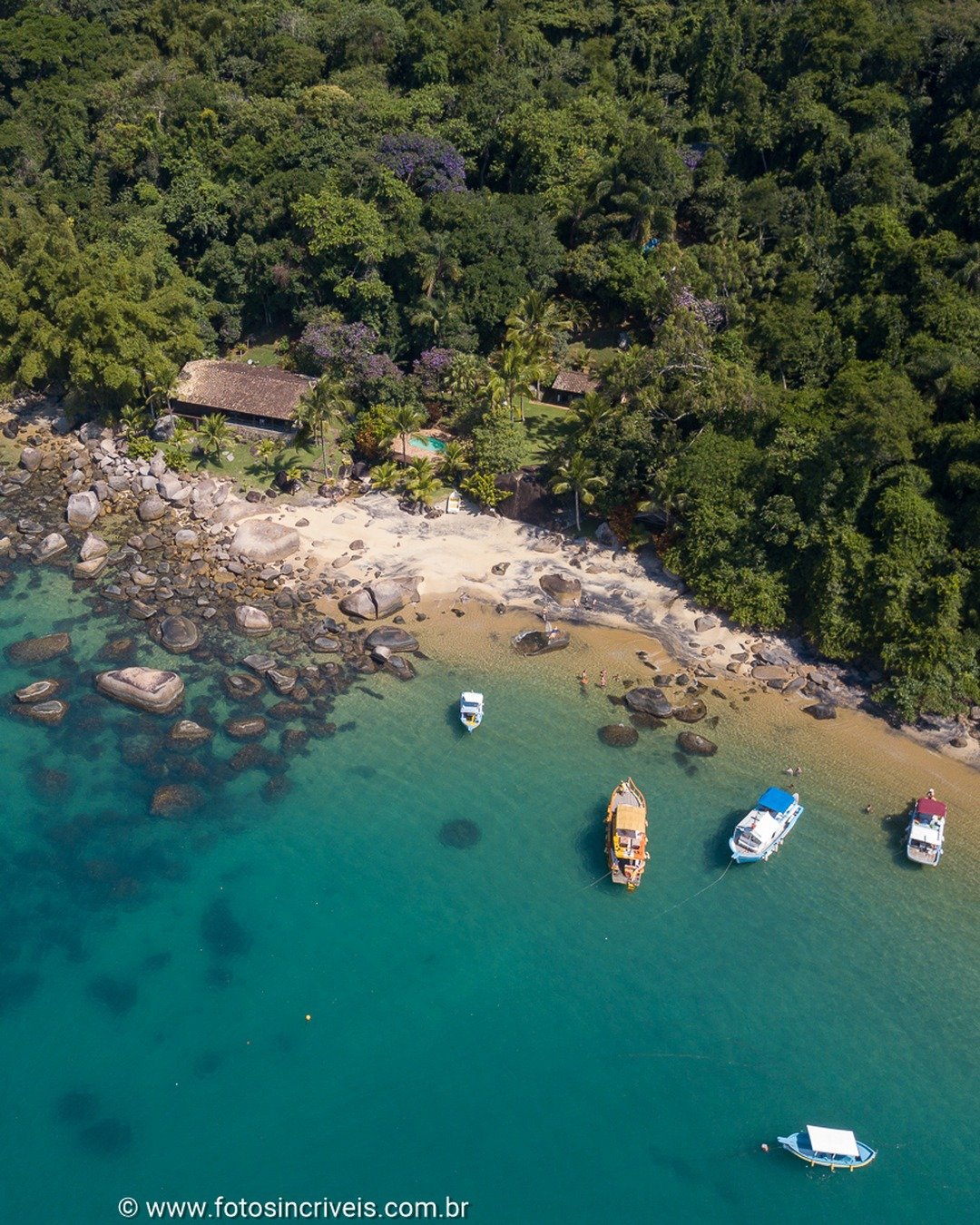 Vista aérea de uma enseada de águas turquesas com praia de areia e pedras, cercada por mata densa e algumas construções rústicas; embarcações ancoradas próximas à praia.