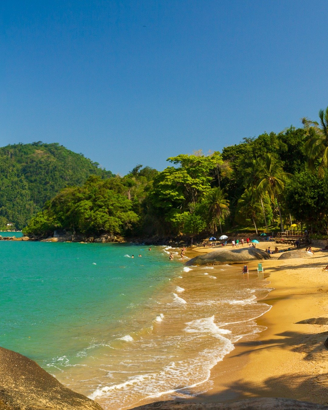Praia de águas turquesa com faixa de areia dourada, grandes rochas graníticas em primeiro plano, coqueiros e mata atlântica à direita e morros verdes ao fundo sob céu azul limpo.