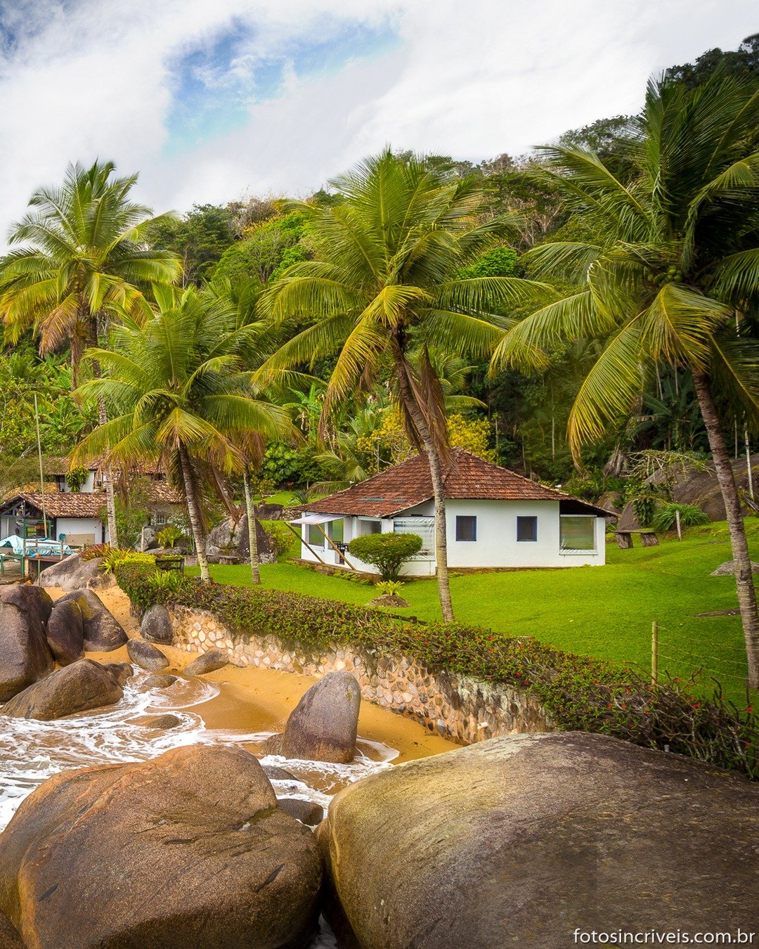 Praia de águas claras com ondas quebrando entre grandes rochas, coqueiros e vegetação tropical densa, com casas brancas de telha à beira-mar sob céu azul com nuvens.