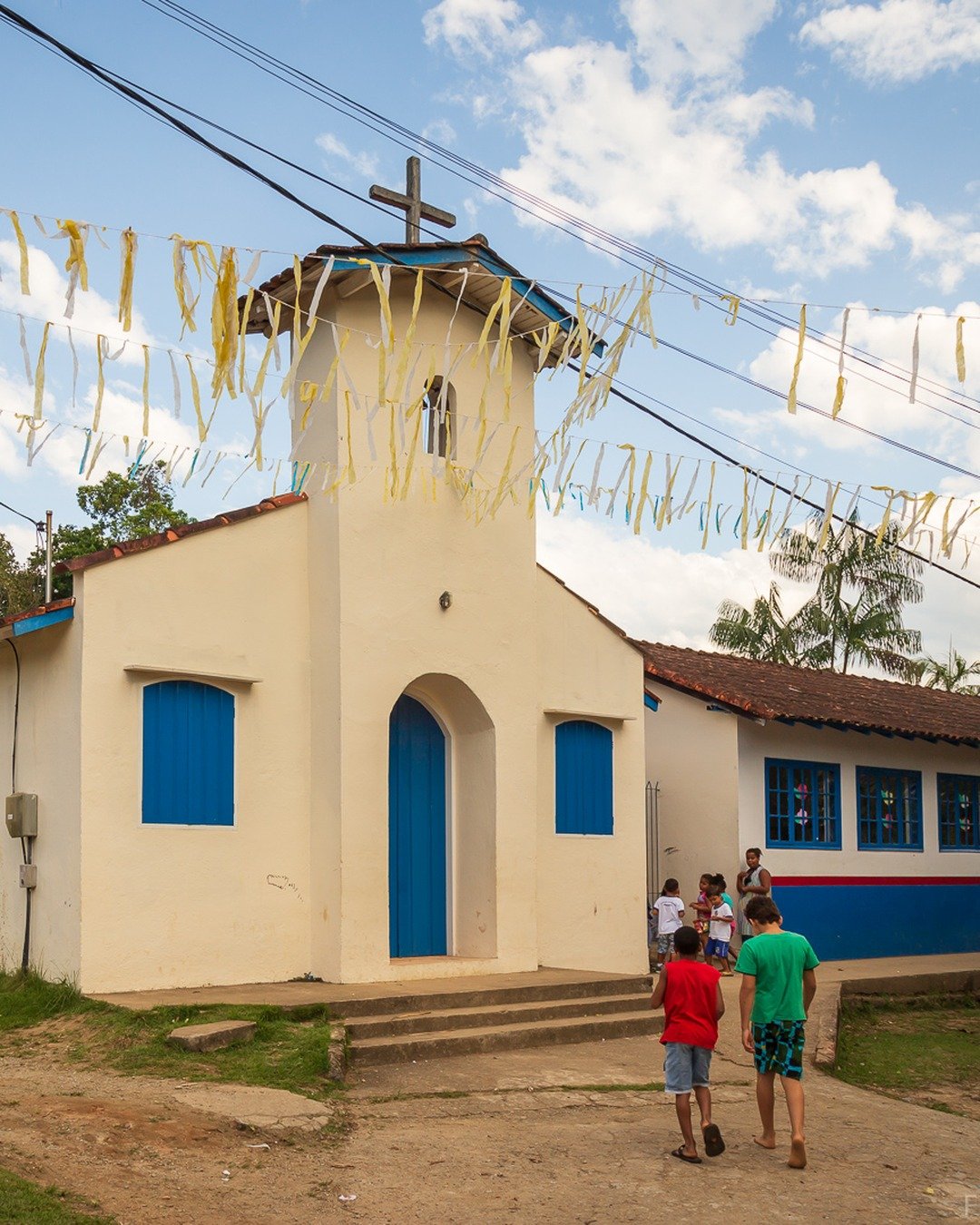 Igreja pequena de fachada branca com portas e janelas azuis e torre com cruz, decorada com bandeirinhas penduradas, sob céu azul com nuvens e morros verdes ao fundo, com pessoas caminhando em primeiro plano.