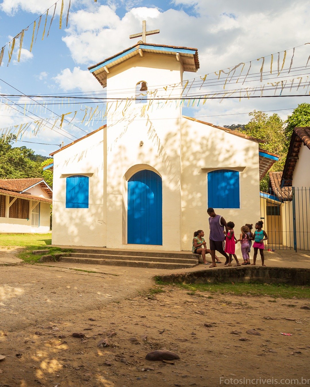 Praça de terra em vila com capela branca de porta e janelas azuis, crianças brincando e bandeirinhas coloridas penduradas sob céu com nuvens.