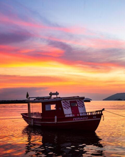 Barco vermelho ancorado em águas calmas ao entardecer, com céu em tons de rosa, laranja e azul e silhueta de morros ao fundo.