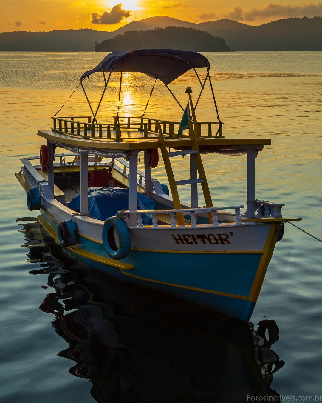 Barco turístico ancorado em águas calmas ao pôr do sol, com montanhas ao fundo e céu dourado refletido na água.