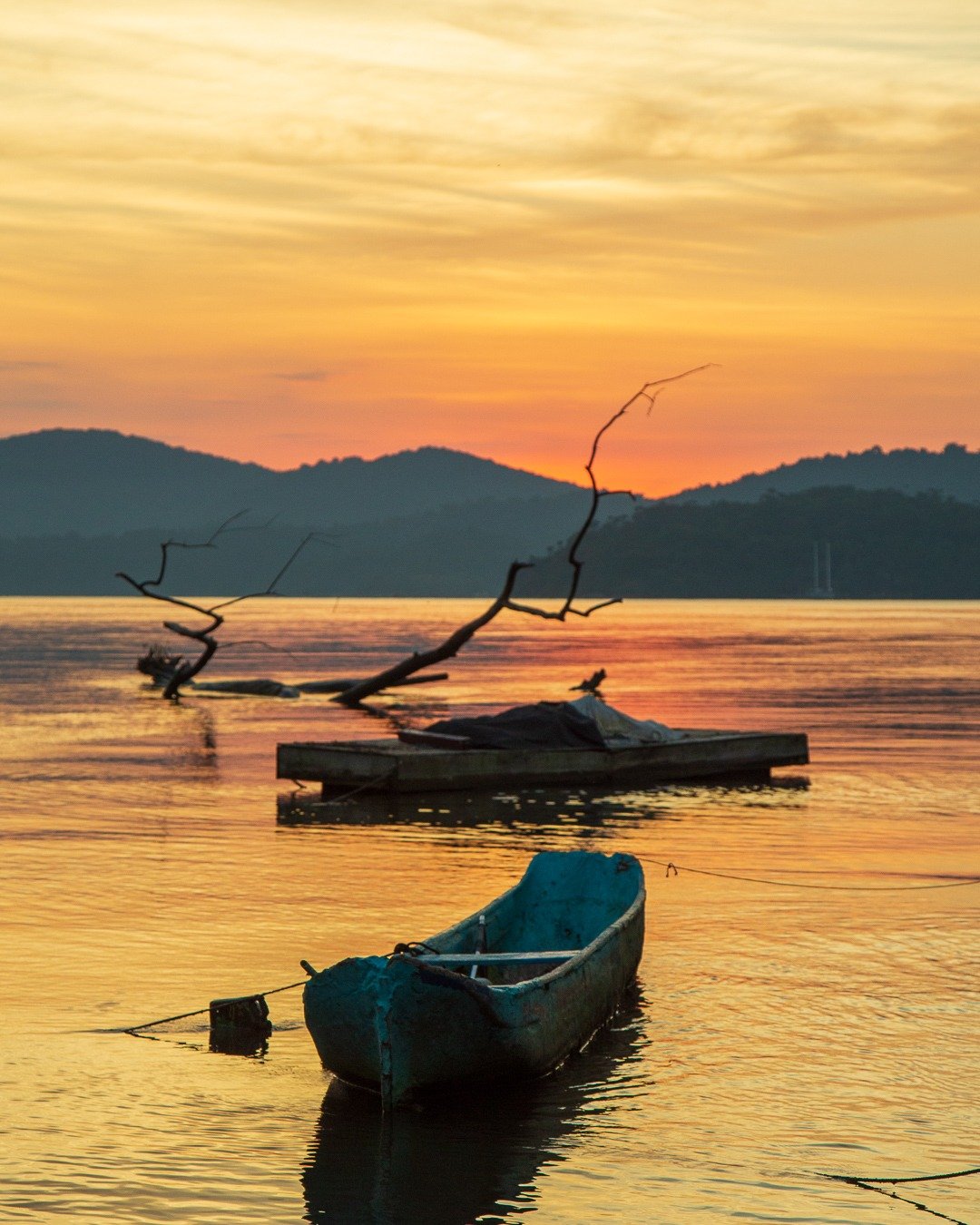 Cena de pôr do sol sobre água calma, com montanhas silhuetadas ao fundo, uma canoa azul em primeiro plano, um galho seco emergindo da água e uma pequena plataforma de madeira com um barco coberto.