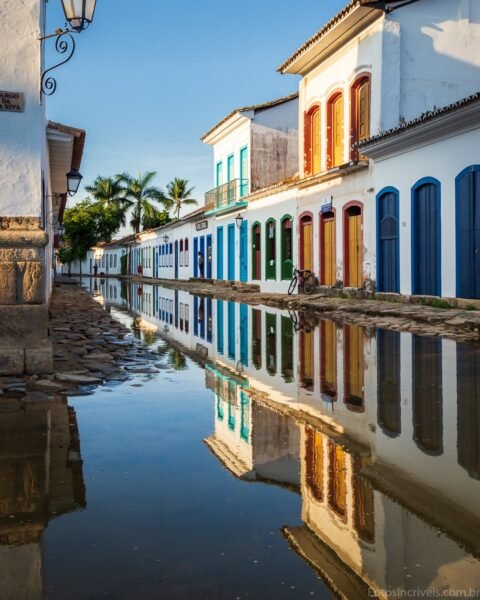 Rua colonial de Paraty alagada, com casarões brancos de portas e janelas coloridas refletidos na água parada sob céu azul.