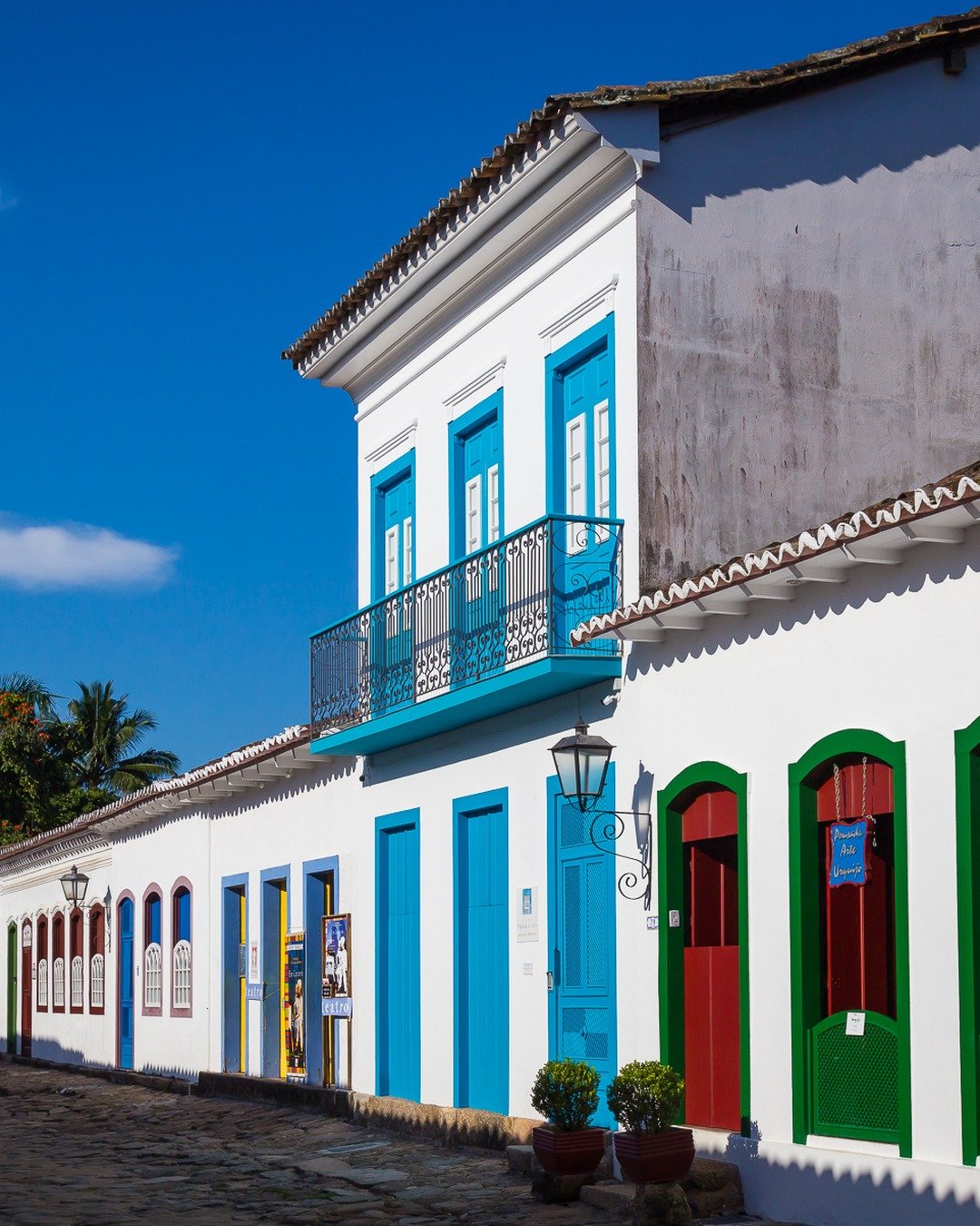 Rua de pedras em centro histórico com casario colonial colorido (portas azuis, verdes, vermelhas e amarelas), palmeiras ao fundo e céu azul com nuvens.
