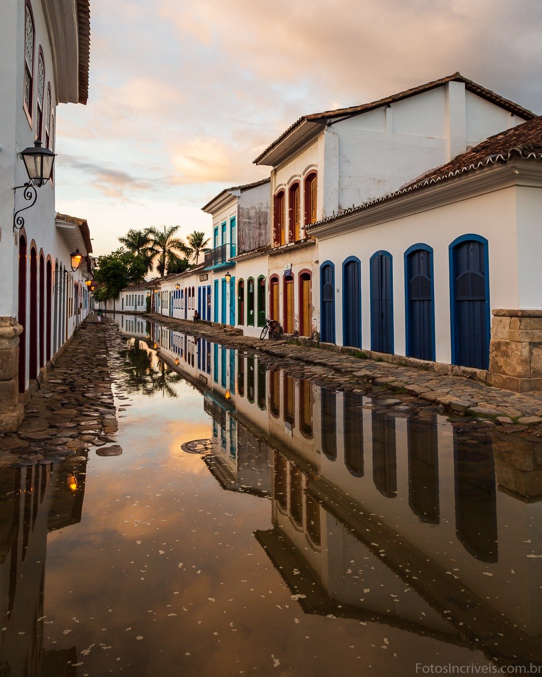 Rua histórica alagada em centro colonial, com casarões de fachadas brancas e portas azuis refletidos na água parada sob céu de fim de tarde.
