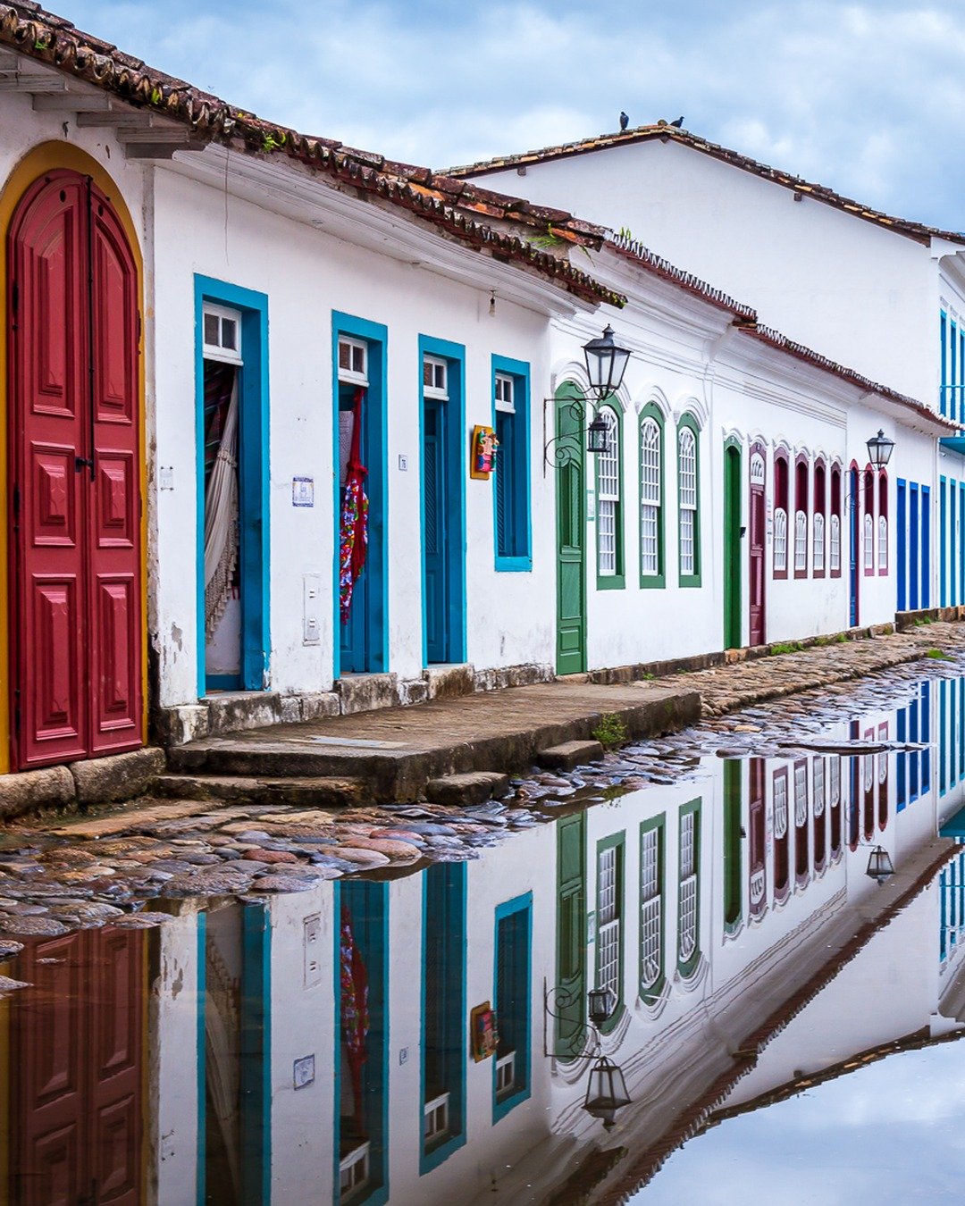 Rua histórica de casario colonial colorido com o chão de pedra alagado, refletindo as fachadas e portas coloridas sob céu nublado, com morro verde ao fundo.