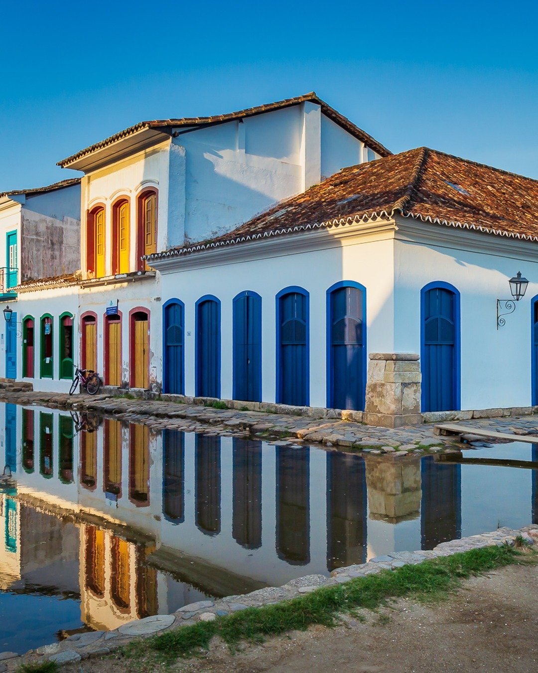 Rua histórica de Paraty com casarões coloniais coloridos, portas e janelas azuis, refletidos na água que cobre a rua encharcada sob céu azul claro.