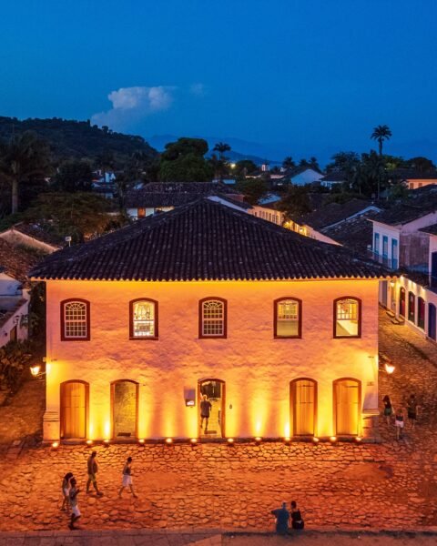 Casarão colonial iluminado por refletores quentes ao anoitecer, com rua de pedra e pessoas caminhando, cercado por telhados e morros ao fundo sob céu azul.
