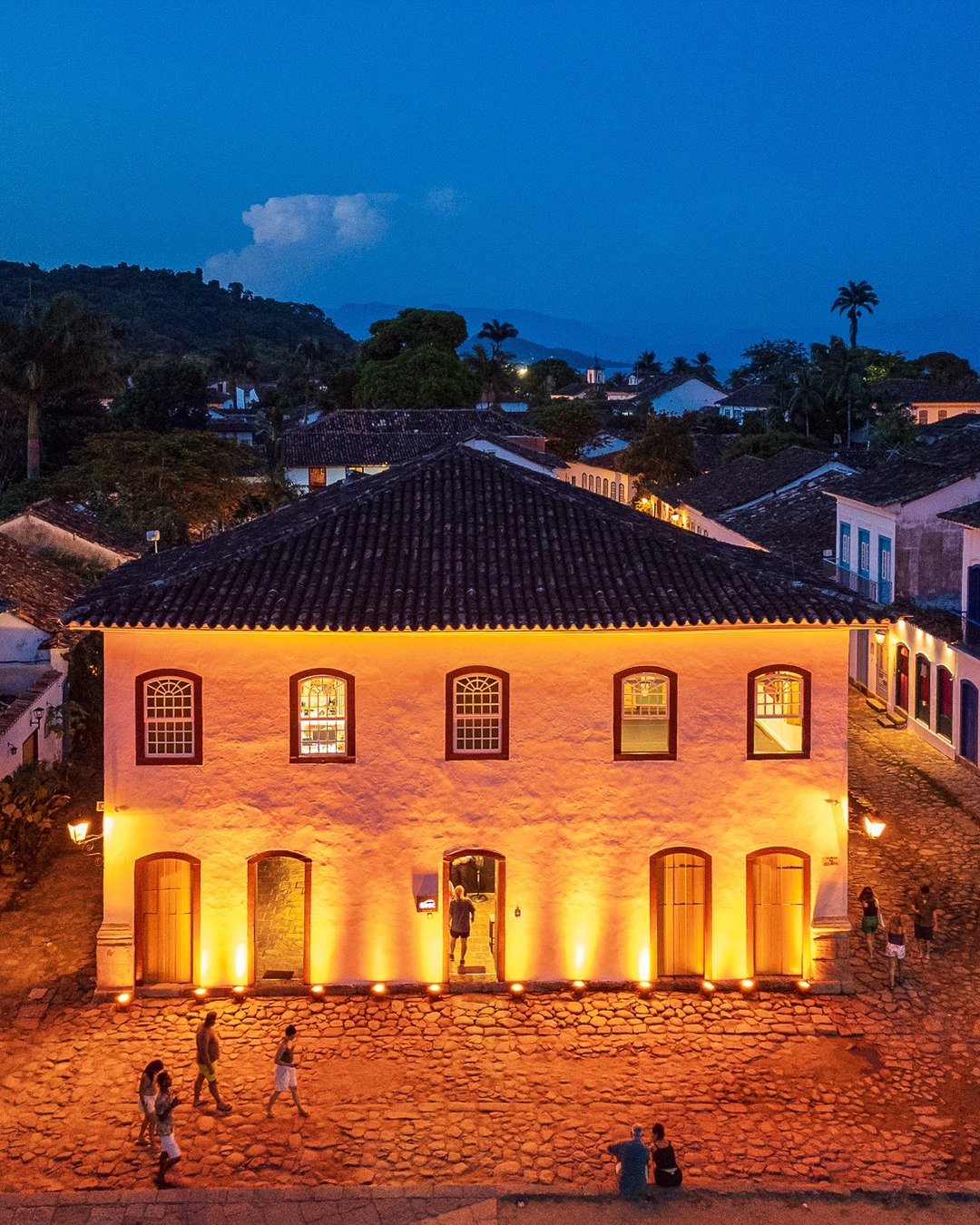 Casarão colonial iluminado por refletores quentes ao anoitecer, com rua de pedra e pessoas caminhando, cercado por telhados e morros ao fundo sob céu azul.