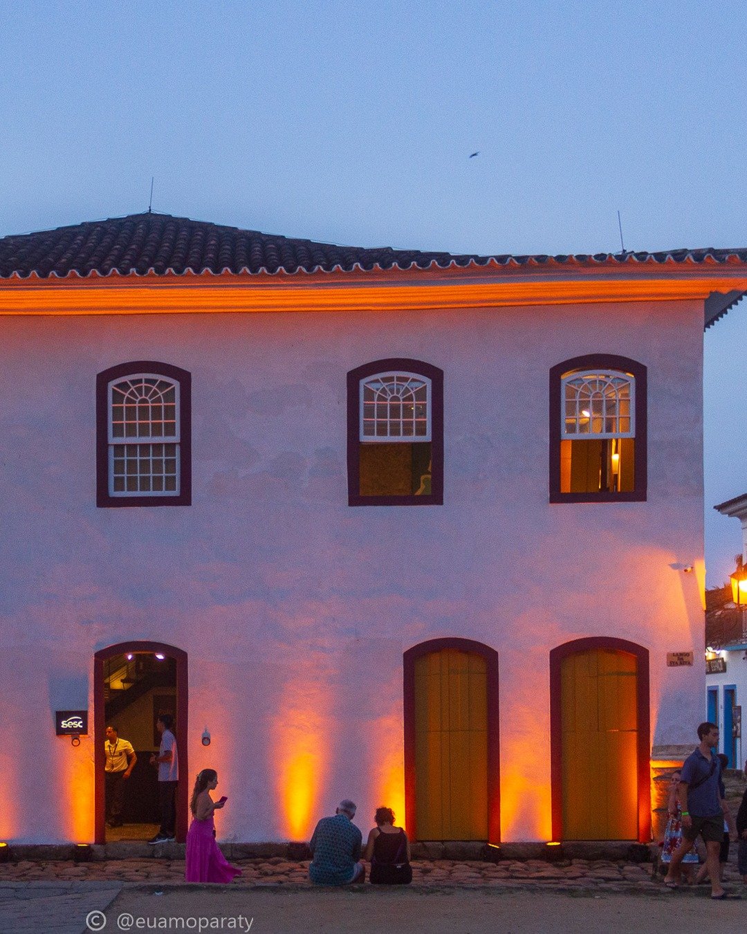 Prédio histórico colonial branco, de dois andares, com janelas e portas em arco, iluminado por refletores laranja ao anoitecer; pessoas circulam na calçada de pedra em frente.