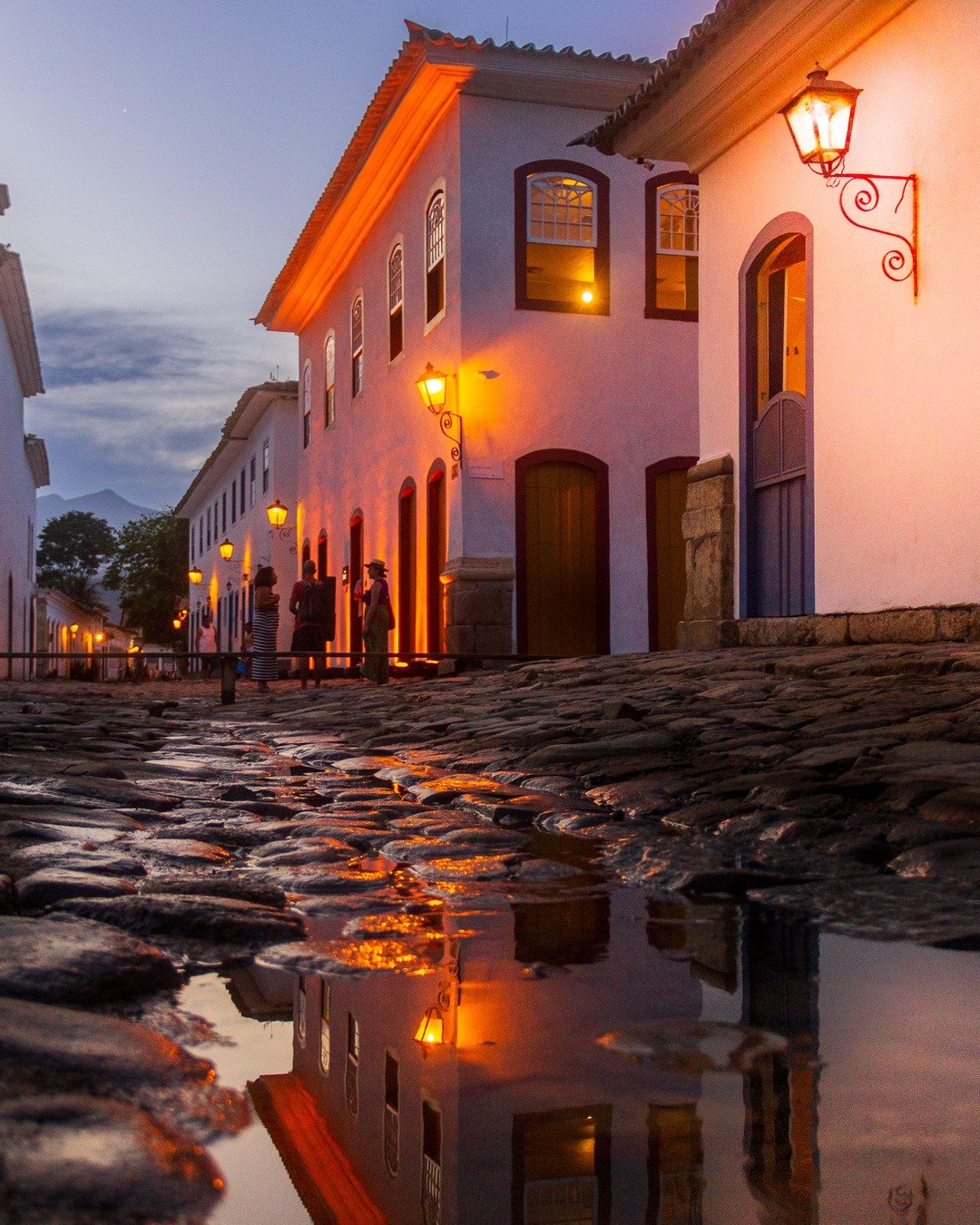 Rua histórica de pedras com casarões coloniais iluminados por lanternas ao anoitecer, com reflexo em poça d'água no primeiro plano.