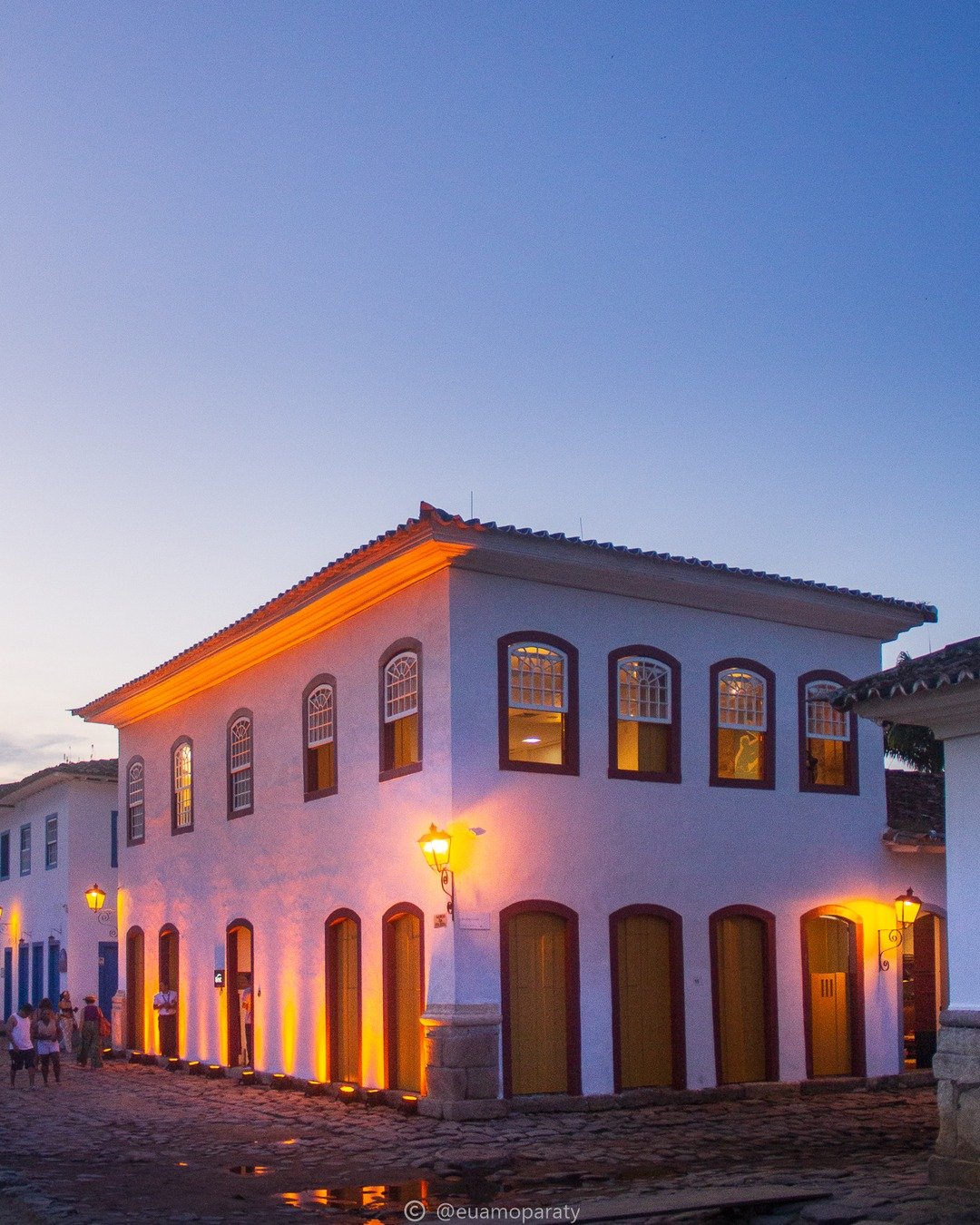 Prédio colonial branco de dois pavimentos iluminado por luzes quentes ao crepúsculo, com rua de pedras e poste de luz em primeiro plano.