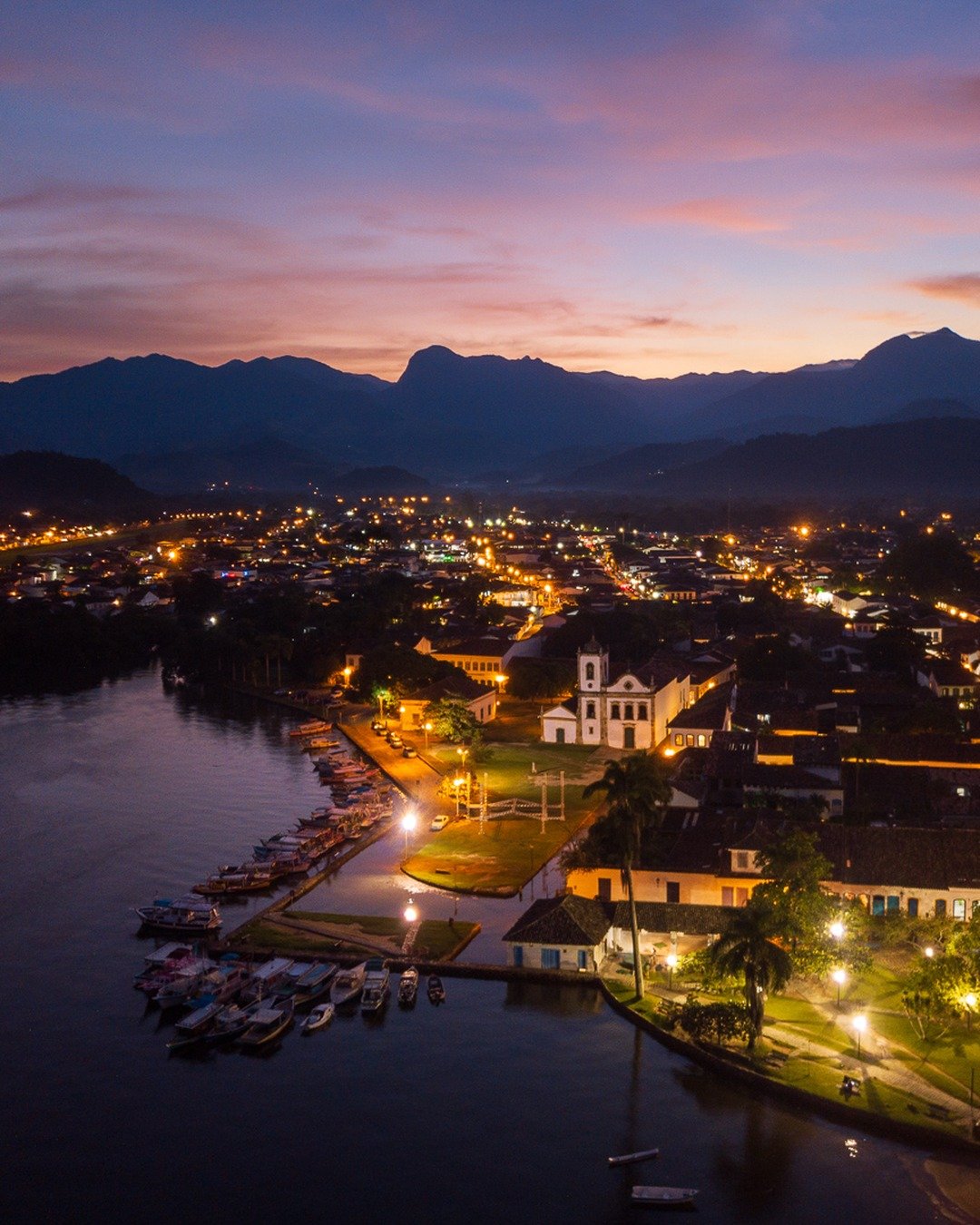 Vista aérea noturna do centro histórico à beira d'água, com a igreja iluminada, cais com barcos e montanhas ao fundo.