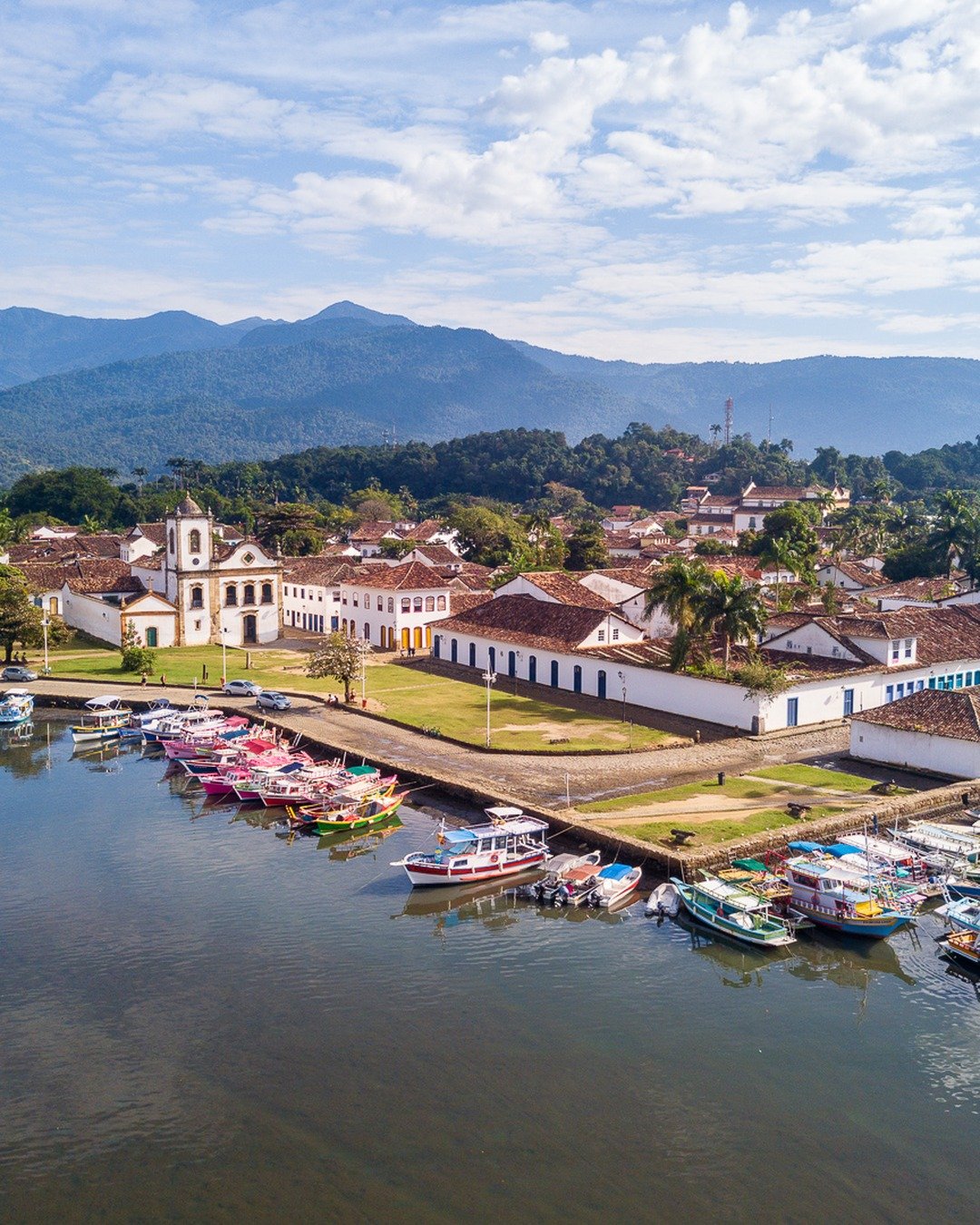 Vista aérea do centro histórico à beira-mar, com a igreja, casarios coloniais, barcos ancorados e montanhas ao fundo.