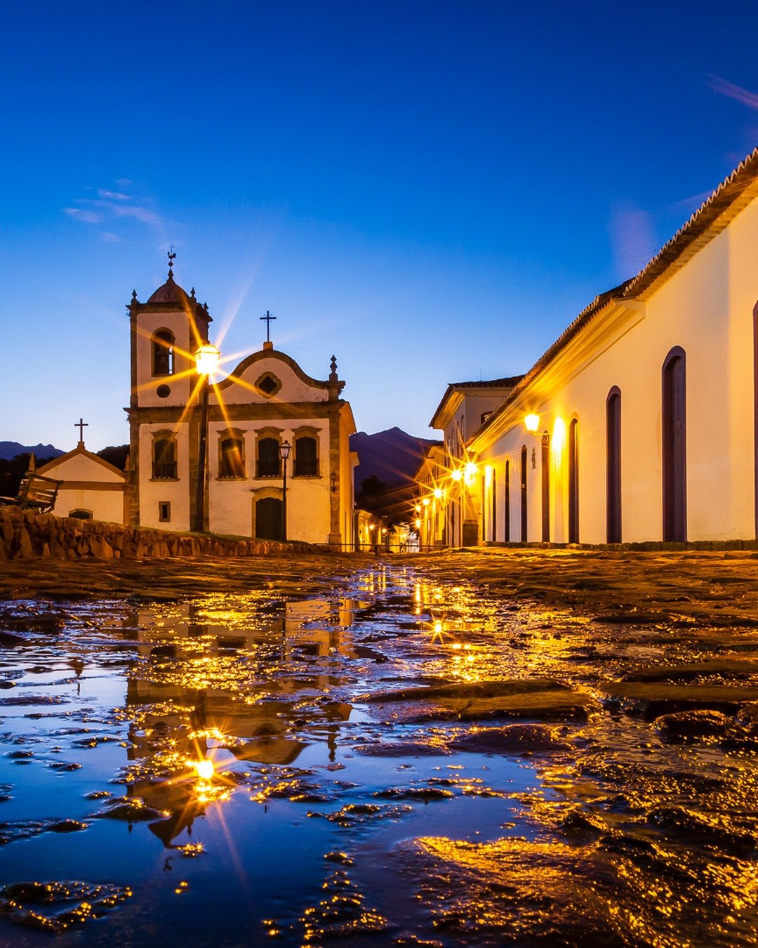 Rua histórica de pedra molhada à noite, com a igreja ao fundo, casario colonial iluminado e reflexos no chão.