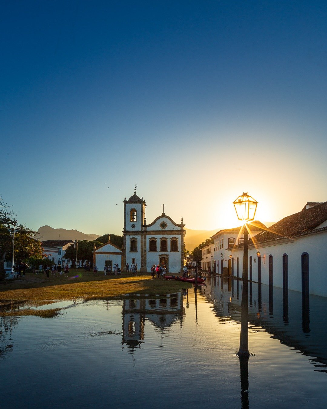 Igreja colonial branca no centro histórico, com rua alagada refletindo o céu dourado do pôr do sol.