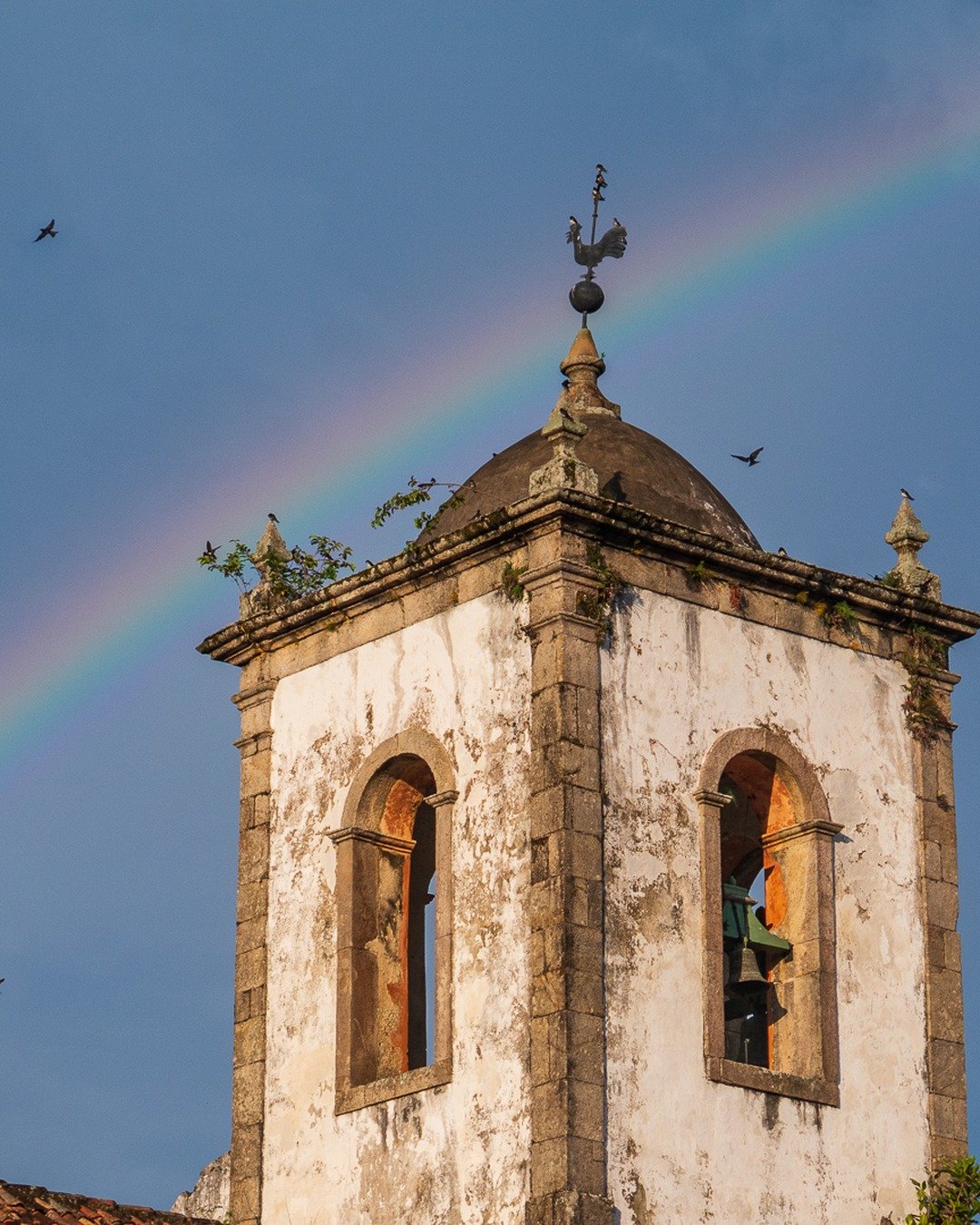 Torre da igreja com sino e cúpula encimada por cata-vento de galo, sob céu azul cortado por arco-íris e pássaros em voo.